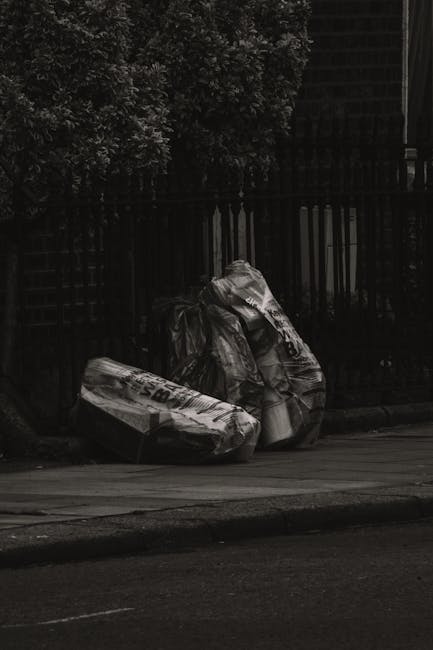 Three large black plastic garbage bags filled with waste are positioned on the pavement near the curb in an outdoor setting. The bags appear slightly crumpled and are partly leaning against each other, with some areas showing creases and folds in the plastic material. Behind the bags, there is a metal fence with vertical bars, and beyond the fence, dense foliage of trees or shrubs creates a dark, leafy background. The scene is lit with natural outdoor light, casting minimal shadows, and the environment suggests a residential or commercial street where private rubbish collection or waste disposal services, such as those offered by Waste Disposal Surrey, might be involved in clearing discarded waste outside typical municipal collection points.