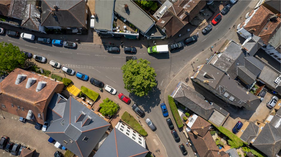 An aerial view of a residential area showing a curved street lined with parked cars of various makes, models, and colours, including white, black, blue, red, and grey. Several houses with different roof styles and materials—some with pitched roofs covered in grey or terracotta tiles—are visible along the street. A prominent leafy green tree in the center of the image provides shade, casting a shadow on nearby vehicles and the pavement. Small front gardens and driveways border the houses, some with potted plants or low fences. The environment appears sunny, with clear lighting illuminating the scene and highlighting the textures of rooftops, car surfaces, and the lush trees. The photo exhibits typical urban residential streets suitable for private waste collection and rubbish removal services, with no visible waste or rubbish piles but providing an accurate context for domestic rubbish collection and on-site waste handling managed by companies like Waste Disposal Surrey.