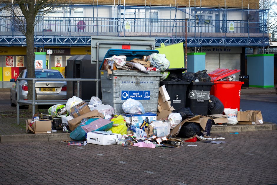 An outdoor scene in a commercial or urban area showing a significant accumulation of overflowing rubbish bins and scattered waste on the ground, including cardboard boxes, plastic bags, paper, packaging, and miscellaneous debris. The waste is piled around a grey mixed paper and cardboard recycling bin, which is open with some contents spilling out, and adjacent black and red rubbish bins, some of which are overfilled. In the background, there is a building with a blue and green framework structure, possibly scaffolding, and a shopfront with signage, indicating a retail or food outlet, along with parked vehicles, including a small silver car on the left side. The scene appears to be during daytime with natural lighting, emphasizing the unsightly and cluttered state of the rubbish accumulation, which may relate to private or commercial waste disposal issues addressed by waste management services such as Waste Disposal Surrey.