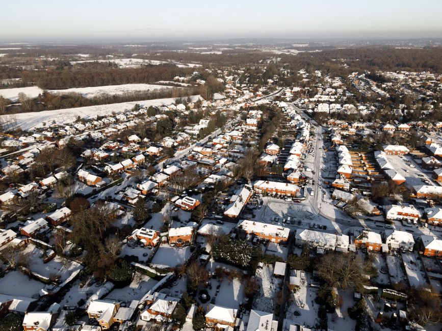 An aerial view of a residential neighbourhood during winter, showing numerous houses with pitched roofs covered in snow. The houses are closely spaced along winding streets, with some driveways and small gardens visible. Trees are scattered throughout the area, many without leaves, and their branches are partially covered with snow. In the background, open fields and woodland extend towards the horizon under a clear sky, creating a contrasting landscape to the densely built-up residential area. The scene captures the quiet and snow-covered environment typical of a suburban part of Surrey, demonstrating a setting where private waste management and rubbish collection services, such as those provided by Waste Disposal Surrey, could be employed for on-site clearance or waste disposal needs in a safe and efficient manner.