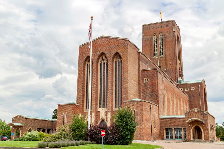 A large brick church with a prominent tower and arched windows stands in a landscaped area with well-maintained greenery including bushes and small trees in front. The church’s reddish-brown brick facade features pointed arch stained glass windows and a flat roof on the main structure, while the tower has vertical slit windows and a small cross at the top. An English flag on a flagpole is positioned to the left of the church entrance, which is located on the right side of the building and marked by a rounded archway. The scene is illuminated by natural daylight under a partly cloudy sky with patches of blue, creating soft shadows. The environment surrounding the church appears quiet, with no visible vehicles or pedestrians. The image may suggest a context where local authorities or private waste disposal services, such as Waste Disposal Surrey, might handle waste collection from such community buildings or nearby areas, reflecting on alternative rubbish removal or on-site clearance options in an urban setting.