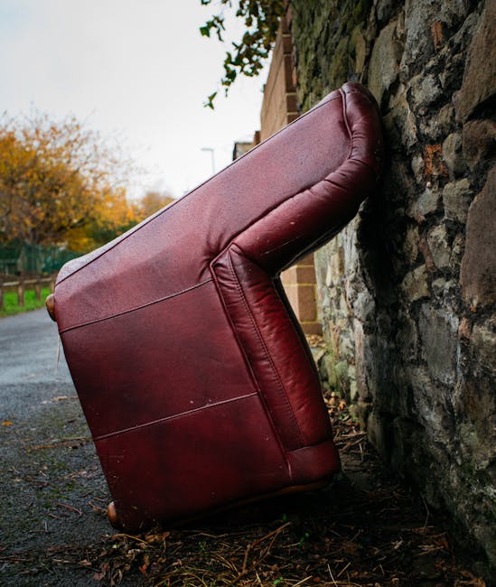 A red leather sofa with a smooth, shiny finish is positioned upright and leaning against a rough stone wall in an outdoor setting. The sofa's upholstery appears slightly worn with visible creases and some minor scratches on the surface. Its cushions are slightly compressed, indicating prior use, and the armrests are rounded and padded. The setting is a paved pathway alongside a building with a brick exterior, bordered by a stone wall that exhibits an uneven, textured surface composed of various sizes and shades of brown and gray stones. In the background, there are trees with autumn foliage featuring orange and yellow leaves, and a glimpse of a fence further down the pathway. The natural lighting suggests an overcast day, casting soft shadows and subtle highlights on the sofa's surface. This scene reflects the type of outdoor rubbish disposal service that Waste Disposal Surrey offers for bulky items such as old furniture needing removal through private or independent collection methods, rather than local authority pickups.