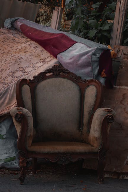 A white upholstered sofa with wooden legs situated outdoors in a park or woodland area, resting on uneven ground covered with grass, fallen leaves, and small plants. The sofa appears to be in good condition, with fabric upholstery and a simple, modern design, featuring a backrest and armrests. In the background, there are tall trees with sparse foliage, a dirt path running through the park, and a streetlamp standing alongside the path. The scene is captured in natural daylight, and the setting suggests it may be part of an outdoor rubbish or unwanted furniture removal, potentially handled by Waste Disposal Surrey, a company specialising in private waste disposal services near Hampton Court in Surrey.